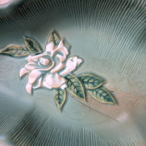 Puede incluir: Un plato de cer&aacute;mica verde con un dise&ntilde;o en relieve de una flor blanca y hojas. El plato tiene un borde festoneado.