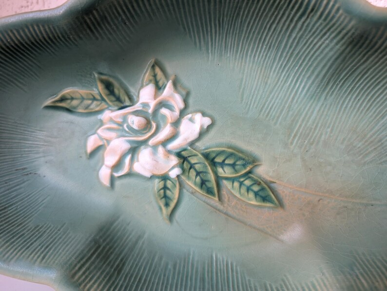 Puede incluir: Un plato de cer&aacute;mica verde con un dise&ntilde;o en relieve de una flor blanca y hojas. El plato tiene un borde festoneado.