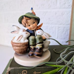 May include: A ceramic figurine of a young person in a green hat and dark blue jacket, surrounded by white rabbits. One rabbit sits in a woven basket. The figurine is on a round, off-white base.