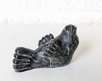 May include: A black soapstone carving of a seal lying on its back with its flippers in the air. The carving is detailed and realistic, with visible fur and wrinkles.