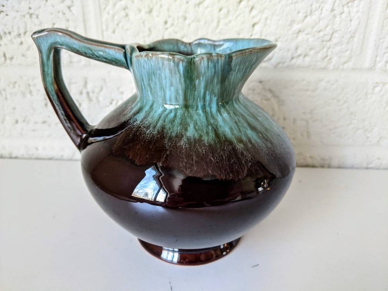 May include: A brown and blue glazed ceramic pitcher with a flared lip and a rounded body. The pitcher has a handle and a base.