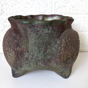 May include: A green and brown ceramic planter with a textured surface and a wide, rounded opening. The planter has three legs and a slightly flared rim.