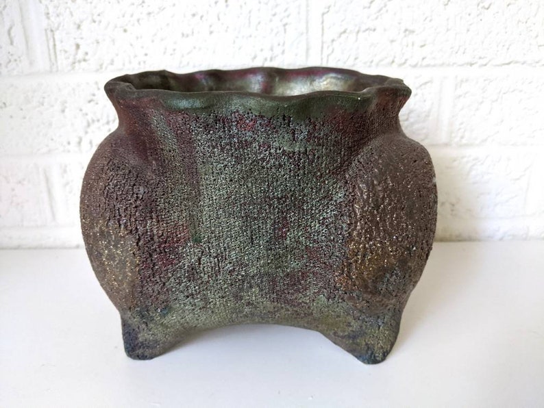 May include: A green and brown ceramic planter with a textured surface and a wide, rounded opening. The planter has three legs and a slightly flared rim.