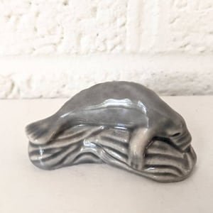 Peut inclure: Une figurine en céramique grise d'un phoque couché sur un lit d'algues. La figurine est détaillée avec des traits réalistes et une finition lisse et brillante.