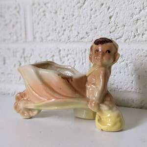 Op de afbeelding: Een keramiek beeldje van een kind dat een kruiwagen met plantenbak duwt. Het beeldje is geschilderd in tinten bruin, geel en roze. De kruiwagen heeft een gele basis en een roze en witte plantenbak.