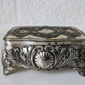 May include: A silver trinket box with an ornate design. The box has a square top with a raised heart-shaped design and a base with intricate floral and shell motifs.