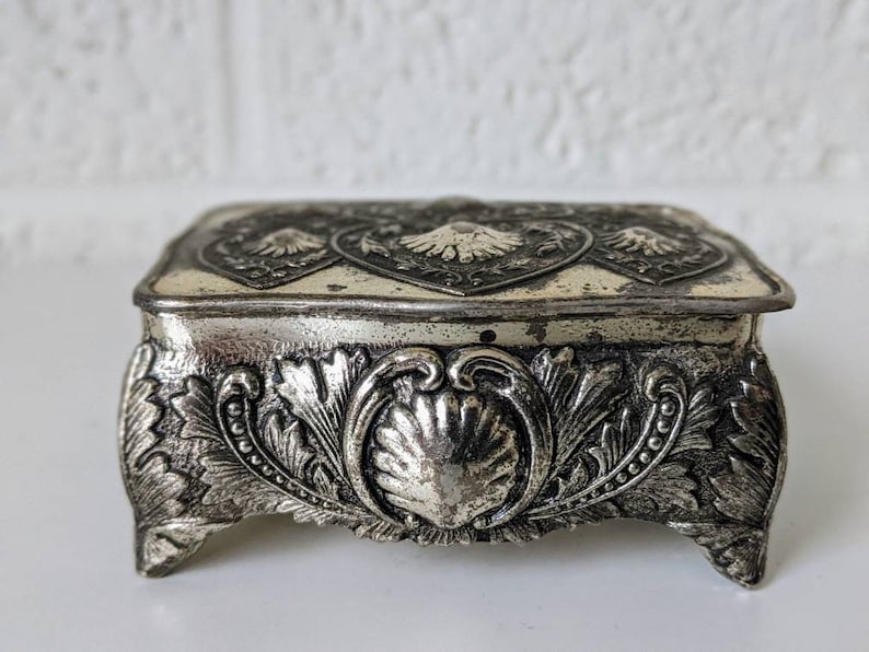 May include: A silver trinket box with an ornate design. The box has a square top with a raised heart-shaped design and a base with intricate floral and shell motifs.