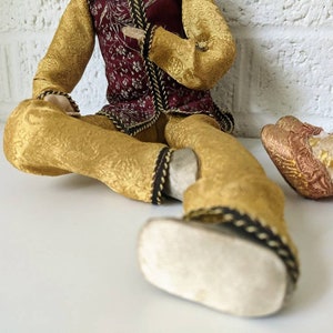 Op de afbeelding: Twee decoratieve poppen gekleed in sierlijke, goudkleurige stof met ingewikkelde patronen. E&eacute;n pop draagt een bordeauxrood vest met gouden accenten. De kleding van de poppen is afgewerkt met zwarte en gouden details. De poppen zitten op een wit oppervlak.