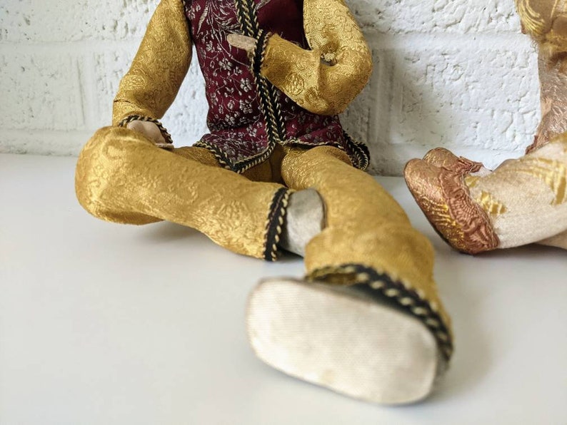 Op de afbeelding: Twee decoratieve poppen gekleed in sierlijke, goudkleurige stof met ingewikkelde patronen. E&eacute;n pop draagt een bordeauxrood vest met gouden accenten. De kleding van de poppen is afgewerkt met zwarte en gouden details. De poppen zitten op een wit oppervlak.