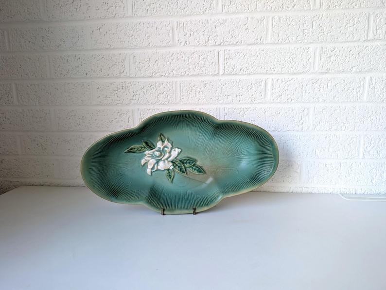 Puede incluir: Un plato de servir de cer&aacute;mica verde con un dise&ntilde;o de flor blanca. El plato tiene un borde ondulado y una superficie texturizada.