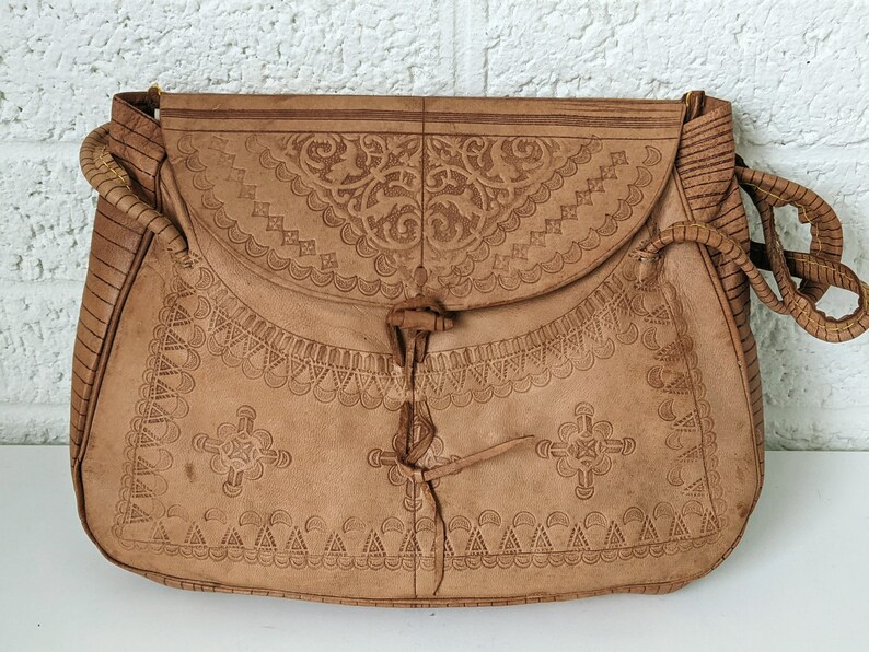 Puede incluir: Un bolso de cuero marr&oacute;n con intrincados dise&ntilde;os en relieve. El bolso tiene un cierre de solapa con una correa de cuero anudada.