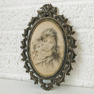 May include: A vintage oval-shaped picture frame with an ornate gold metal design. The frame holds a black and white drawing of a mother and child.