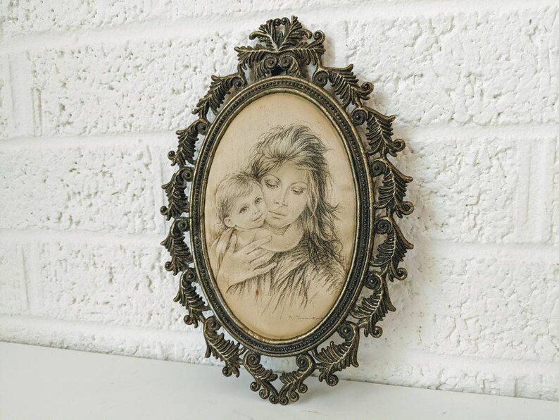 May include: A vintage oval-shaped picture frame with an ornate gold metal design. The frame holds a black and white drawing of a mother and child.