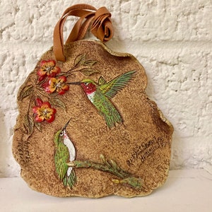 Op de afbeelding: Een keramische wandhanger met twee kolibries en rode bloemen. De hanger is bruin met een beige achtergrond en heeft een leren band om op te hangen.