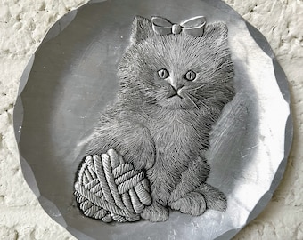 Puede incluir: Un plato de metal plateado con una imagen en relieve de un gatito gris con un lazo rosa, sosteniendo una bola de lana.
