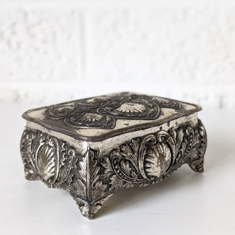 May include: A silver trinket box with intricate floral and shell designs. The box has a square shape with a raised lid and sits on four feet.