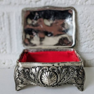 May include: A silver trinket box with an ornate design and a red velvet interior. The box has a hinged lid and sits on four feet.