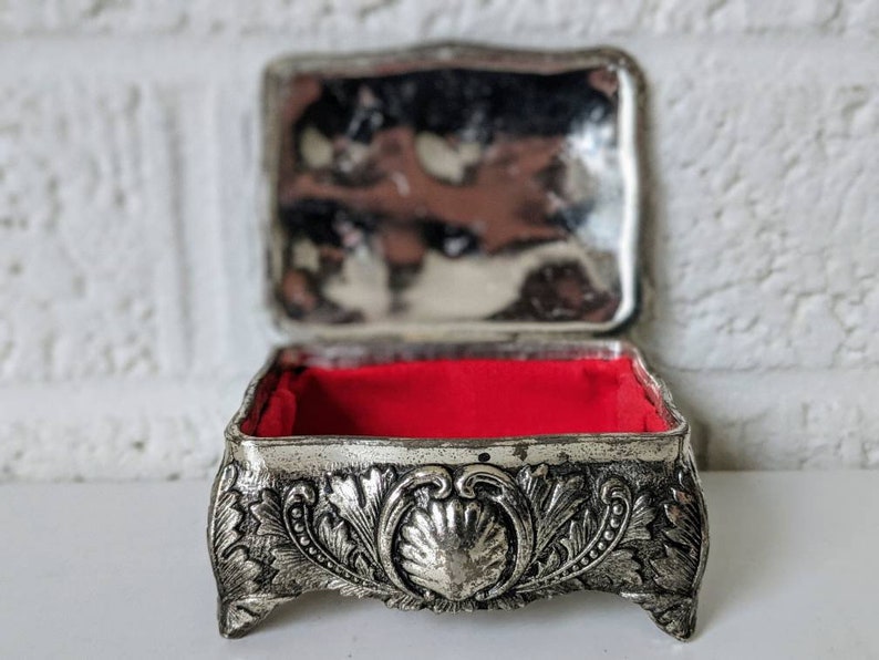 May include: A silver trinket box with an ornate design and a red velvet interior. The box has a hinged lid and sits on four feet.