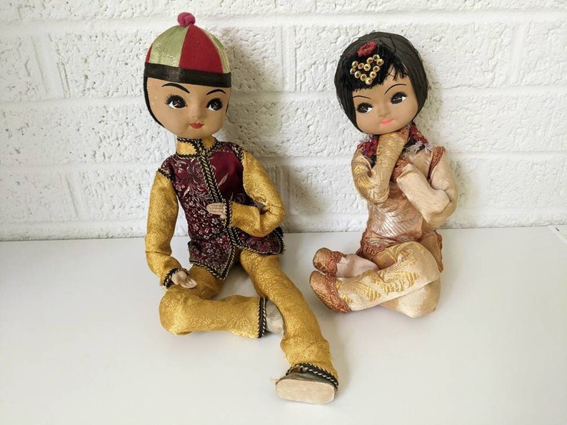 Op de afbeelding: Twee vintage Aziatische poppen. De mannelijke pop draagt een goud- en bordeauxrode outfit met een bijpassende hoed. De vrouwelijke pop draagt een goud-beige outfit met een decoratief hoofddeksel. Beide poppen zitten.