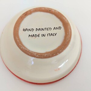 May include: The bottom of a white ceramic dish with a red rim. The dish has the words "Hand Painted and Made in Italy" written on the bottom.