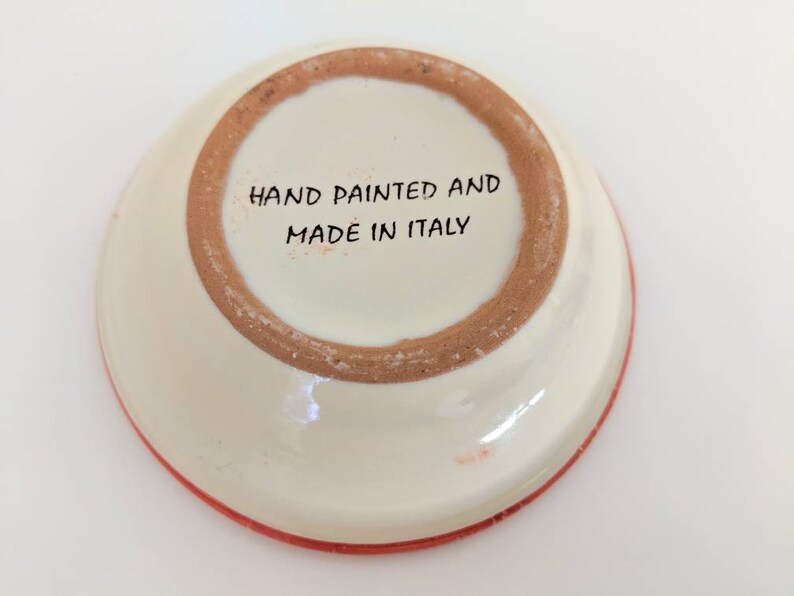 May include: The bottom of a white ceramic dish with a red rim. The dish has the words "Hand Painted and Made in Italy" written on the bottom.
