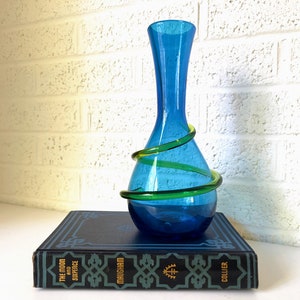 May include: A blue glass vase with a green glass coil wrapped around it. The vase is sitting on a book titled "The Moon and Sixpence" by W. Somerset Maugham.