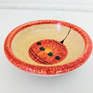 May include: A small, round ceramic bowl with a red rim and a hand-painted orange ornament design. The ornament has black lines and black dots.