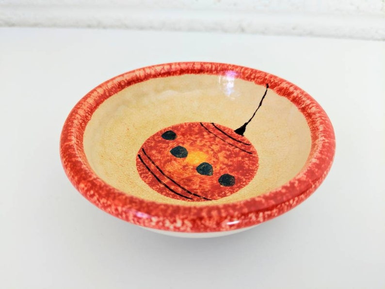 May include: A small, round ceramic bowl with a red rim and a hand-painted orange ornament design. The ornament has black lines and black dots.