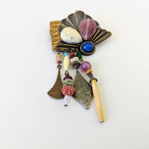 May include: A multi-coloured, beaded and metal brooch with a shell-shaped design. The brooch features a variety of beads, including pink, blue, white and brown. The brooch is attached to a metal pin.