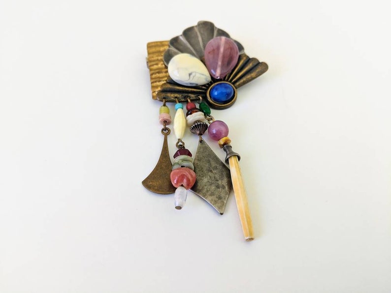 May include: A multi-coloured, beaded and metal brooch with a shell-shaped design. The brooch features a variety of beads, including pink, blue, white and brown. The brooch is attached to a metal pin.