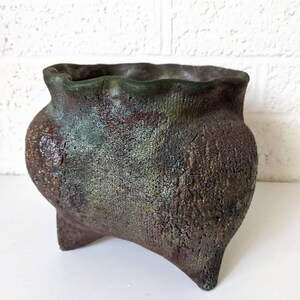 May include: A rustic, green and brown ceramic planter with a wide, uneven rim and three short, sturdy legs. The planter has a textured surface and a mottled, aged appearance.