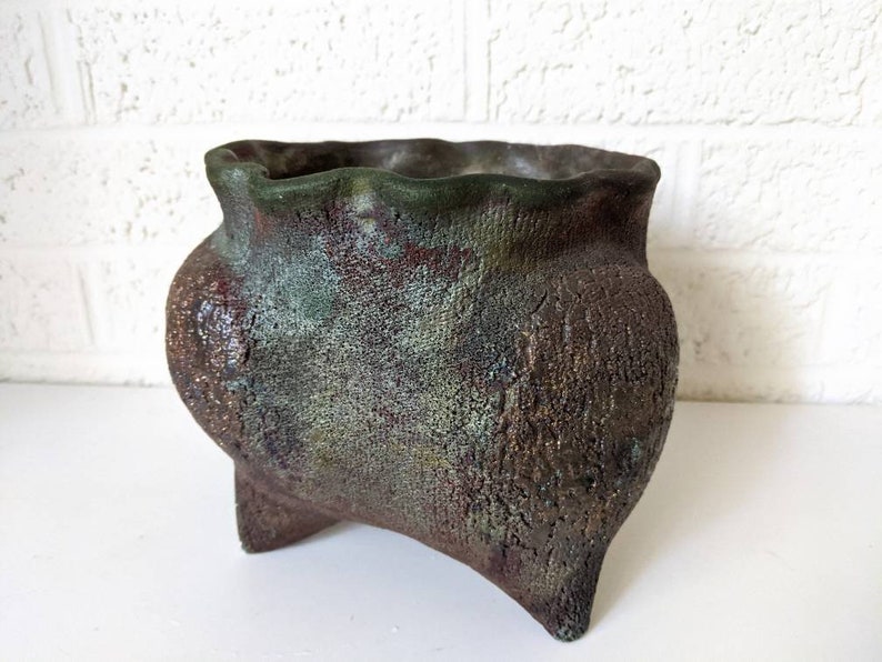 May include: A rustic, green and brown ceramic planter with a wide, uneven rim and three short, sturdy legs. The planter has a textured surface and a mottled, aged appearance.