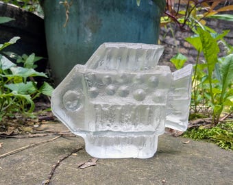 May include: A clear, glass fish sculpture with a textured surface. The fish has a detailed design with fins, a tail, and a round eye. The sculpture is displayed outdoors on a stone surface, with greenery in the background.