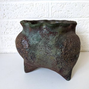 May include: A rustic, green and brown ceramic planter with a wavy rim and three legs. The planter has a textured surface and a unique, hand-crafted look.