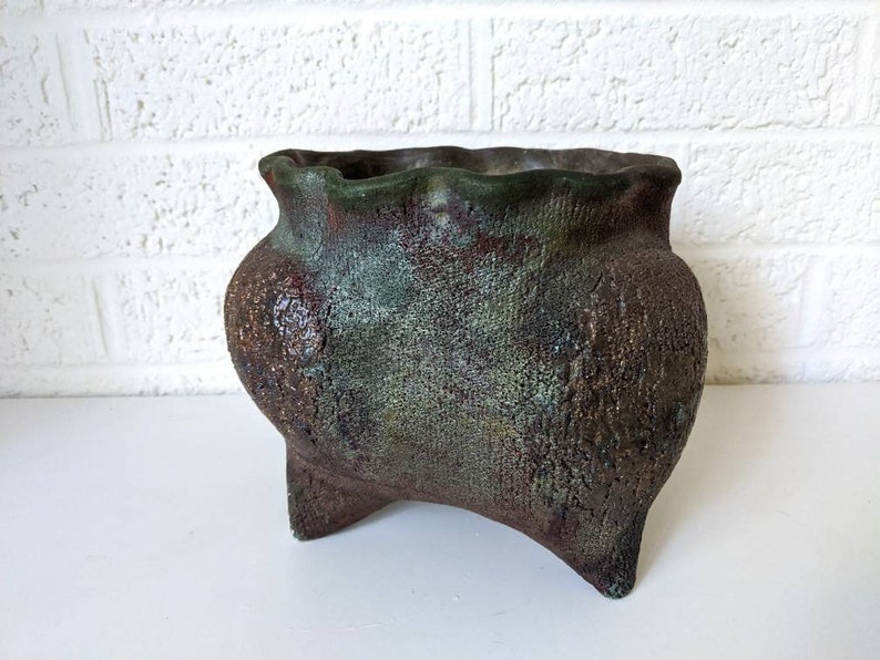 May include: A rustic, green and brown ceramic planter with a wavy rim and three legs. The planter has a textured surface and a unique, hand-crafted look.