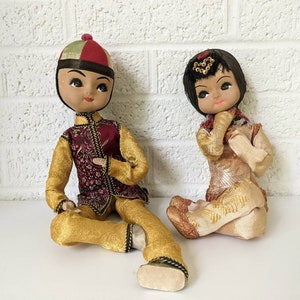 Op de afbeelding: Twee vintage Aziatische poppen, een man en een vrouw, zitten. De mannelijke pop draagt een goud-bordeauxrode outfit met een bijpassende hoed. De vrouwelijke pop draagt een goud-beige outfit met een decoratieve hoofdtooi. Beide poppen hebben beschilderde gezichten.
