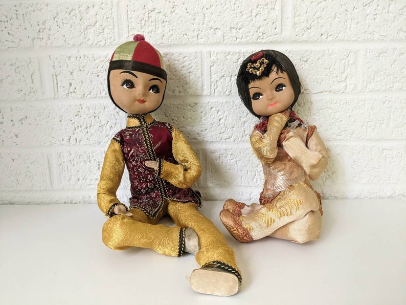 Op de afbeelding: Twee vintage Aziatische poppen, een man en een vrouw, zitten. De mannelijke pop draagt een goud-bordeauxrode outfit met een bijpassende hoed. De vrouwelijke pop draagt een goud-beige outfit met een decoratieve hoofdtooi. Beide poppen hebben beschilderde gezichten.
