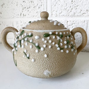 May include: A beige ceramic sugar bowl with a lid, decorated with white and green floral accents. The bowl has a textured surface and two handles.