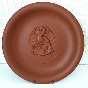 Puede incluir: Un plato de terracota con un diseño en relieve de un dragón sosteniendo un escudo con una llave y una estrella. El plato tiene el texto "Nibelungenstadt 1923" alrededor del escudo.