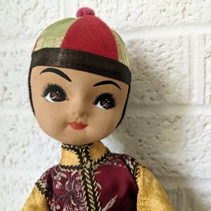 Op de afbeelding: Een vintage pop met een geschilderd gezicht, gekleed in een rode, gouden en groene outfit. De pop heeft een rode en groene hoed met een kleine rode pompon. De outfit van de pop heeft gouden randen en een bordeauxrood vest met bloemenborduurwerk.