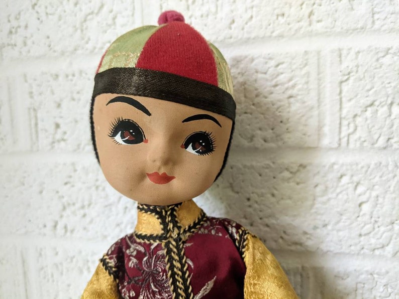 Op de afbeelding: Een vintage pop met een geschilderd gezicht, gekleed in een rode, gouden en groene outfit. De pop heeft een rode en groene hoed met een kleine rode pompon. De outfit van de pop heeft gouden randen en een bordeauxrood vest met bloemenborduurwerk.