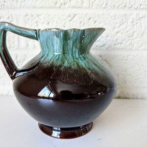 May include: A brown and green glazed ceramic pitcher with a rounded body and a flared lip. The pitcher has a single handle and a glossy finish.