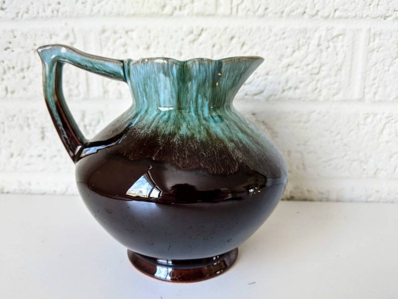 May include: A brown and green glazed ceramic pitcher with a rounded body and a flared lip. The pitcher has a single handle and a glossy finish.