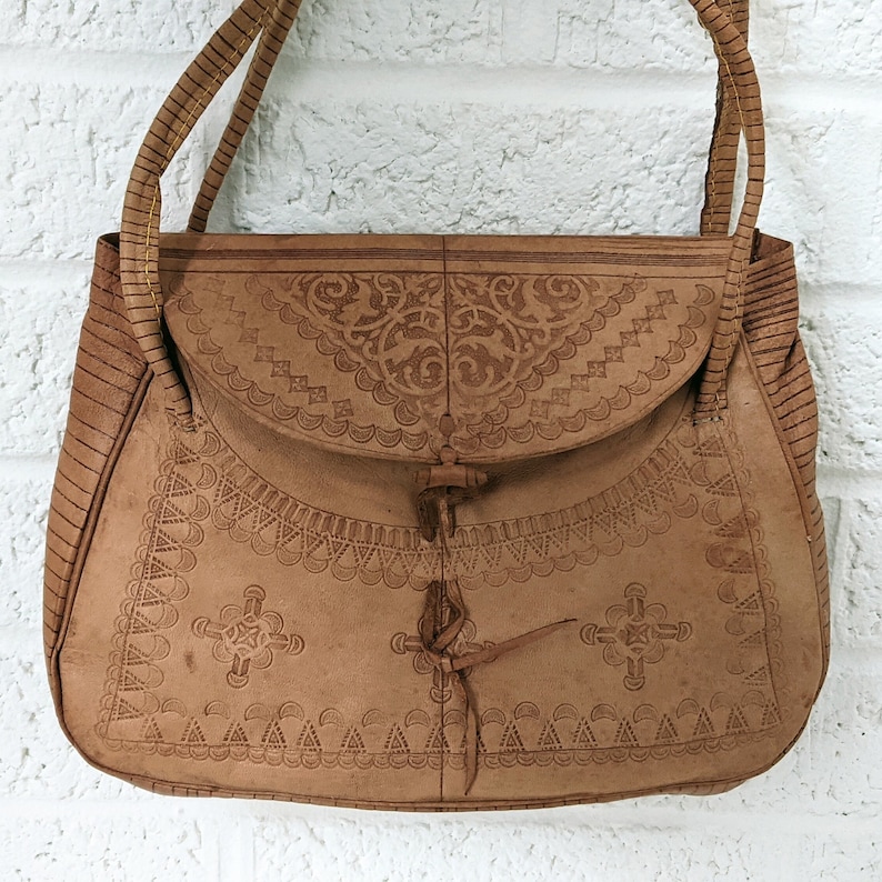 Puede incluir: Un bolso de mano de cuero marr&oacute;n con intrincados dise&ntilde;os en relieve. El bolso tiene un cierre de solapa y una sola correa.