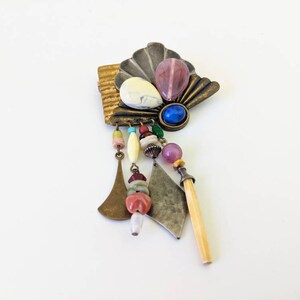 May include: A colourful, multi-layered brooch with a fan-shaped design, featuring a variety of beads, stones, and metal accents. The brooch has a dangling charm with a bone-like piece.
