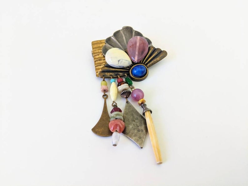 May include: A colourful, multi-layered brooch with a fan-shaped design, featuring a variety of beads, stones, and metal accents. The brooch has a dangling charm with a bone-like piece.