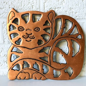 May include: Copper metal cat-shaped trivet with a cutout design. The trivet is a light brown color and has a detailed design of a cat's face and body.