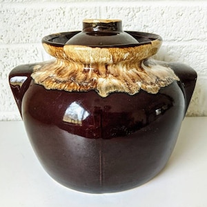 May include: A brown glazed ceramic crock pot with a lid. The crock pot has a white and brown glaze pattern around the top.