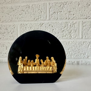 May include: A black circular plaque with a gold-colored depiction of the Last Supper. The plaque is on a white surface.