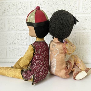 Op de afbeelding: Twee vintage poppen, rug aan rug, gekleed in traditionele Aziatische kleding. De pop links draagt een rood-gouden hoed en een bordeauxrode vest over een gouden outfit. De pop rechts heeft zwart haar en een perzikkleurige outfit met gouden accenten.